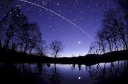 A Discovery űrsikló és a Nemzetközi Űrállomás íve Bakonybélben a Borostyán-kút tavacskája felett a szürkület színeiben. Alattuk a Hold és a Jupiter fénye tükröződik a víz felszínén.